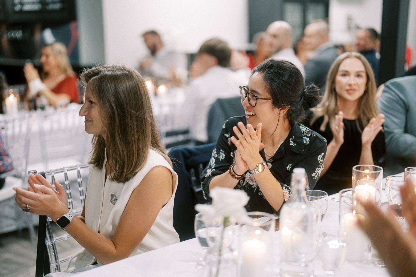 People clapping at an elegant indoor event, seated around a table with candles and glassware, expressing appreciation and enjoyment.