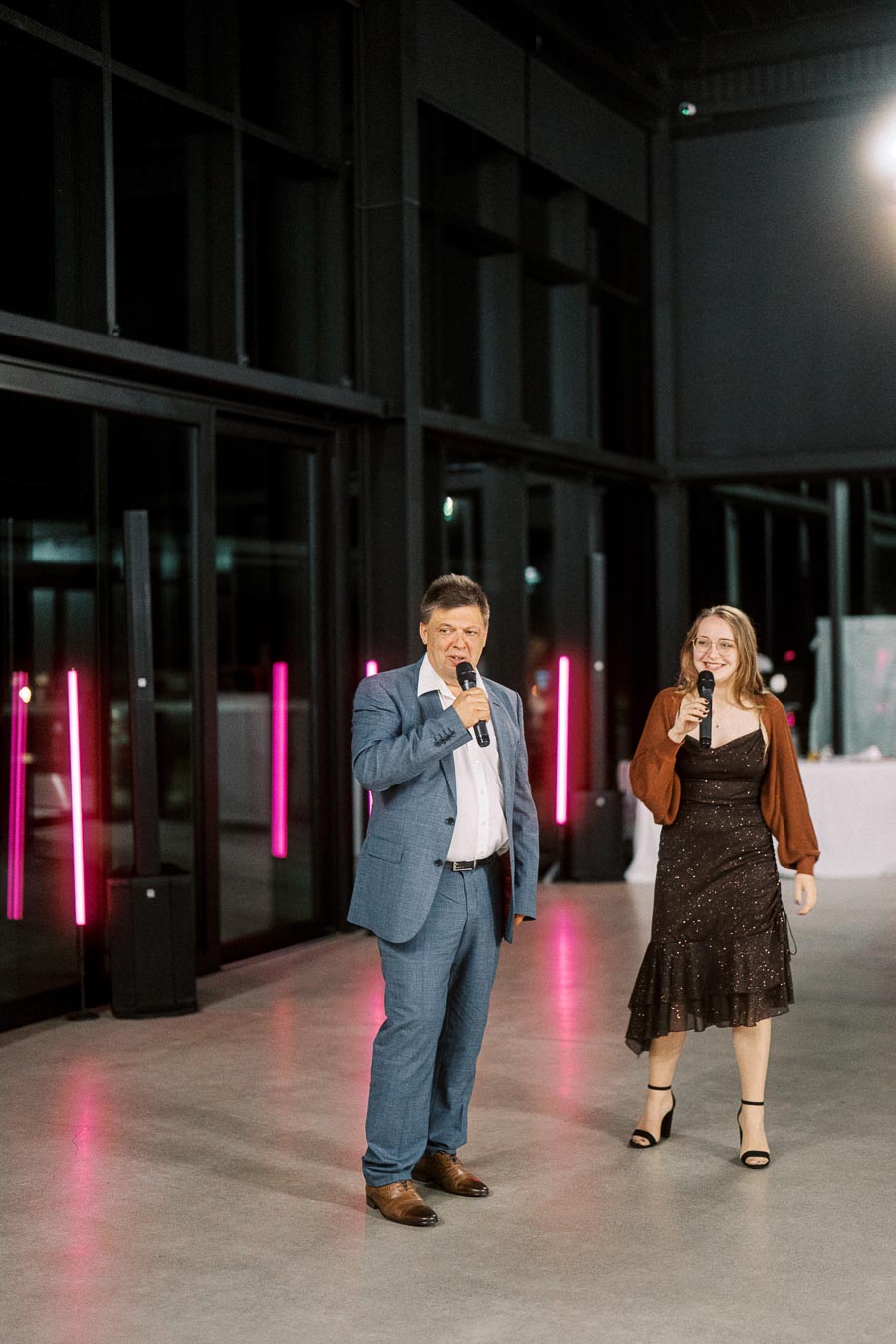 Two people holding microphones stand indoors, dressed formally. The man is in a blue suit, and the woman wears a dark dress with a brown cardigan. They appear to be speaking or singing at an event, with a modern setting and neon pink lights in the background.
