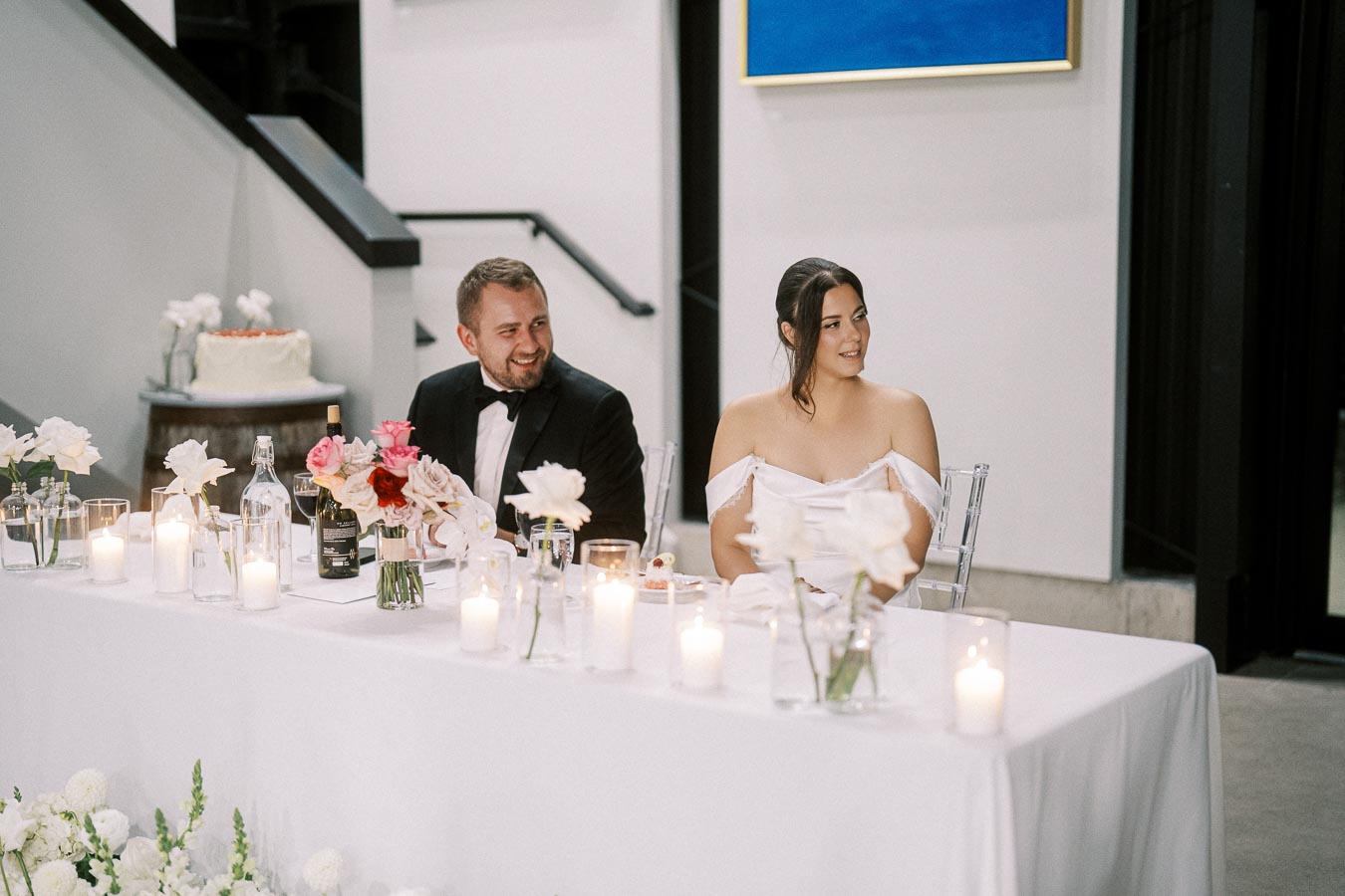 A couple sitting at a decorated wedding table with candles and flowers, smiling during the reception in an elegant venue.
