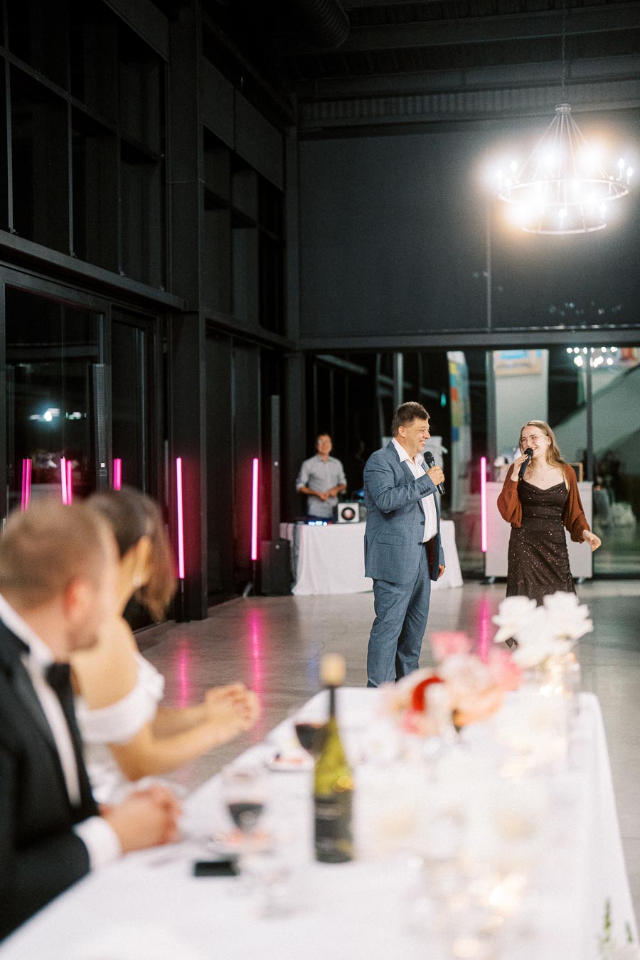 A wedding reception with a man in a suit and a woman in a dress singing into microphones, surrounded by a decorated table, guests in formal attire, and a modern venue with pink neon lights.