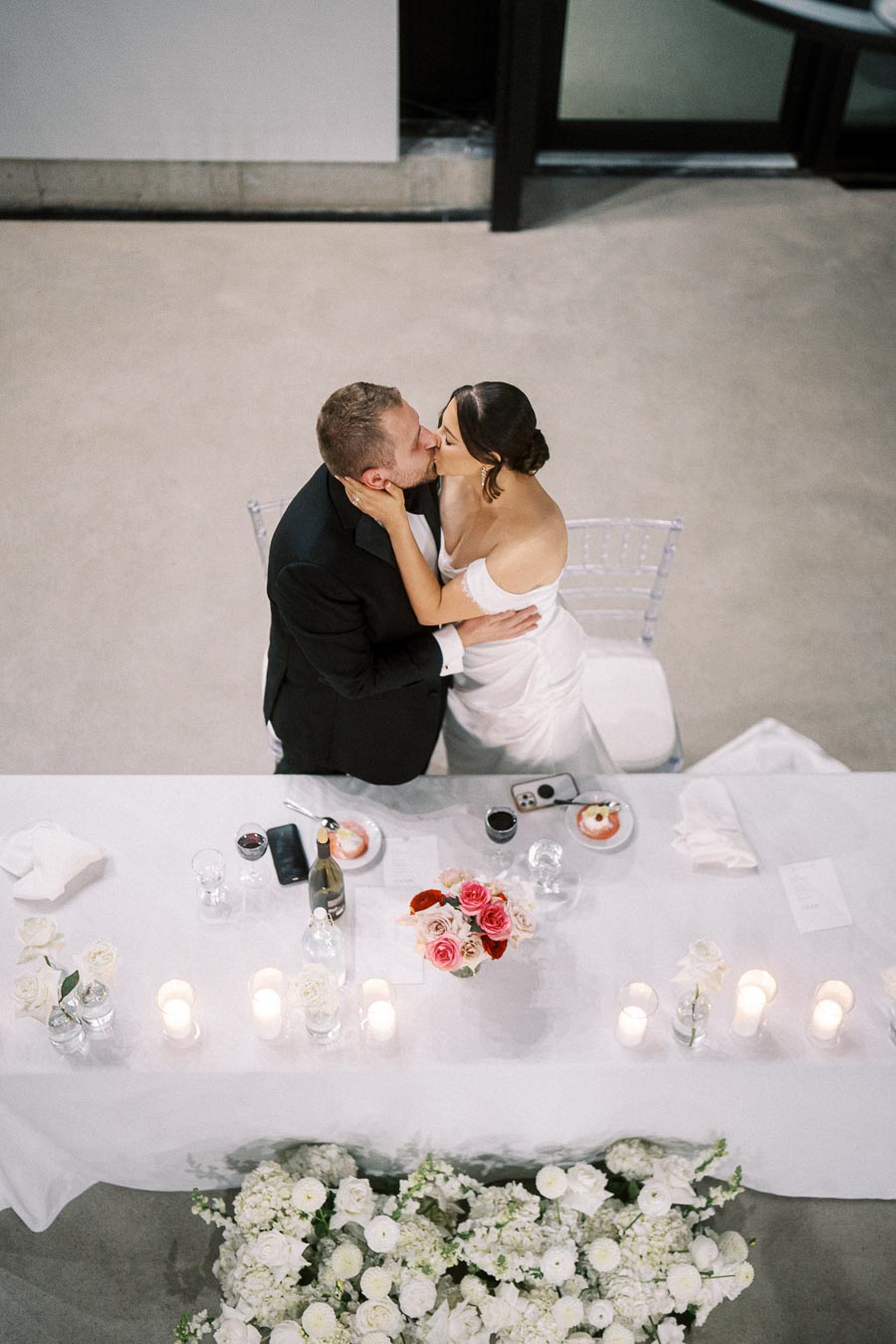 Overhead view of a bride and groom sharing a kiss at their wedding reception table, elegantly decorated with white and pink roses, candles, and glasses, creating a romantic atmosphere.