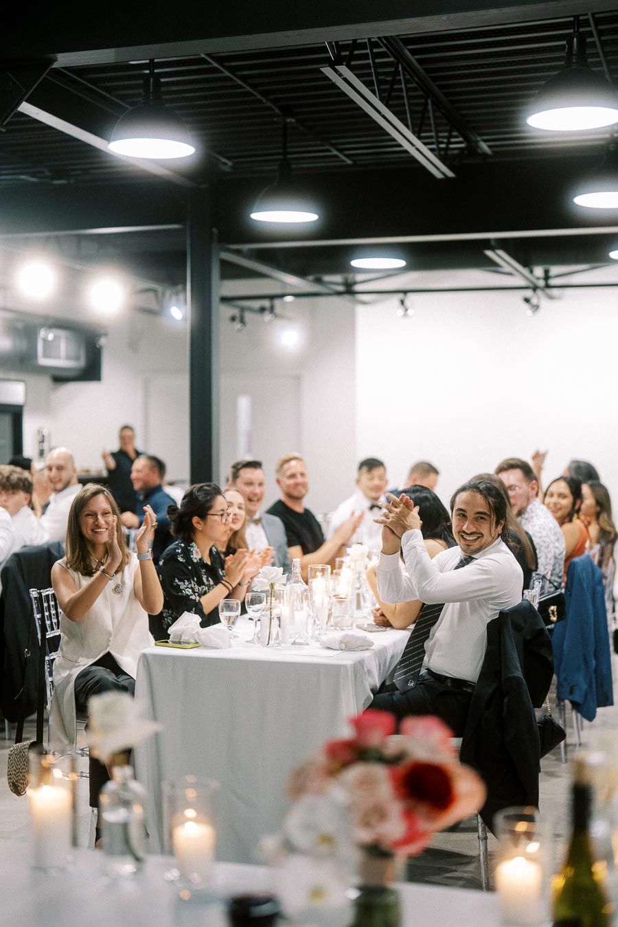 Guests clapping and enjoying a lively atmosphere at a modern indoor event, with tables set elegantly and decorated with candles and flowers.