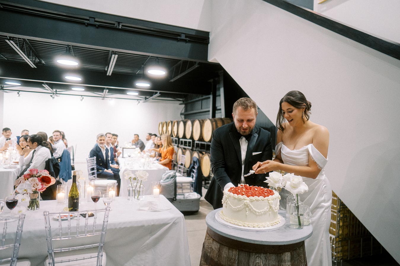 A couple in wedding attire cutting a white frosted cake topped with strawberries, surrounded by guests seated at decorated tables in a modern, airy venue with wine barrels in the background.