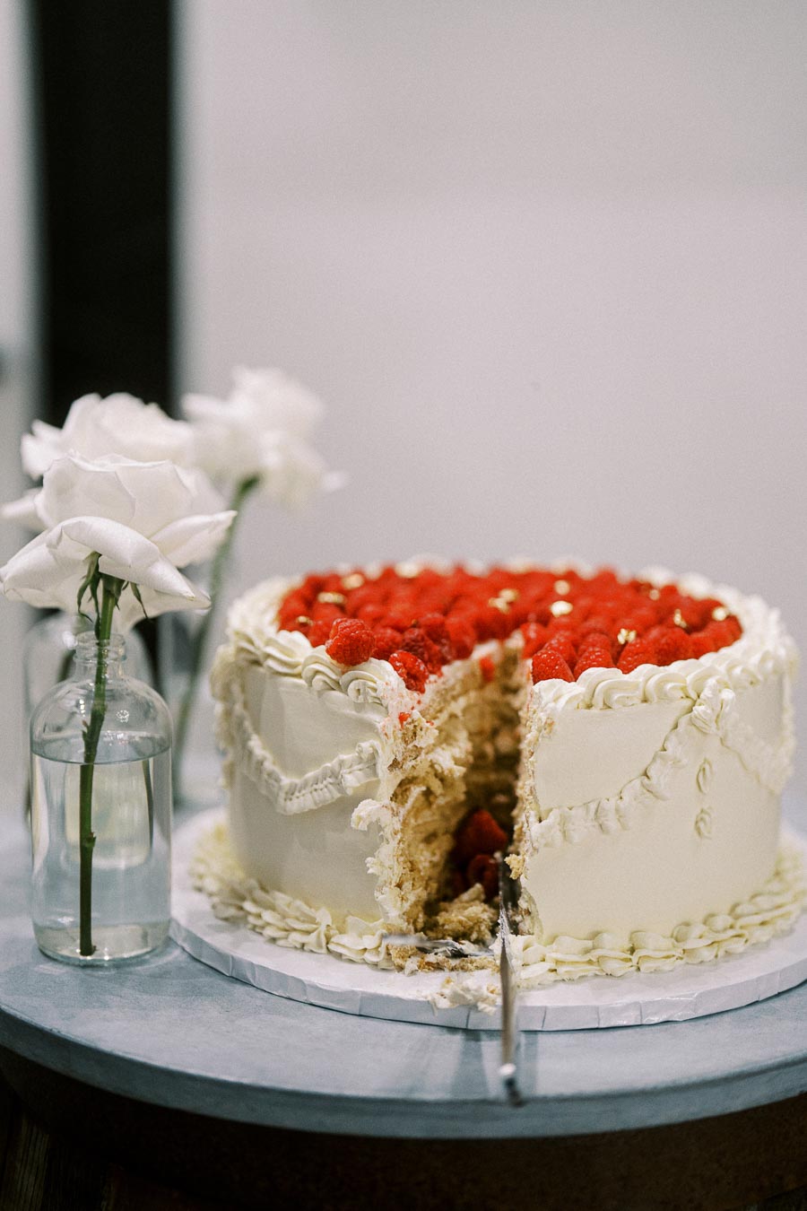 A partially sliced berry-topped wedding cake with decorative white frosting and fresh roses in a glass vase on the table.