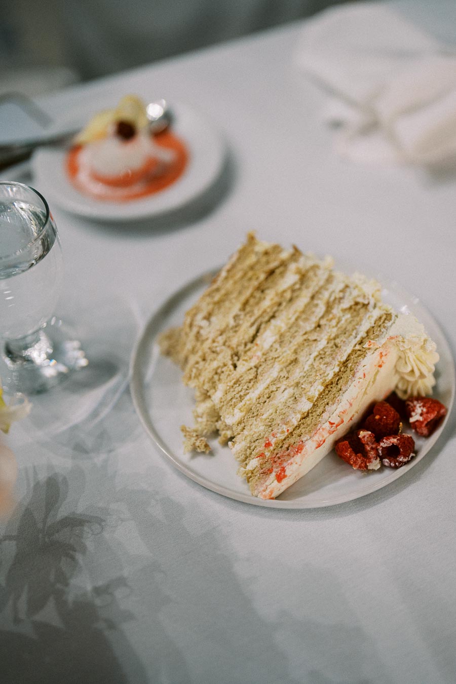 Slice of layered cake garnished with fresh raspberries on a white plate, accompanied by a glass of water and a dessert in strawberry sauce in the background, set on a white tablecloth.