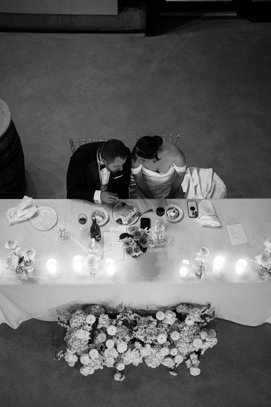 A black-and-white image of a wedding couple seated at an elegantly decorated table adorned with flowers and candles. The bride is wearing an off-the-shoulder gown, and the groom is in a tuxedo. They appear to be sharing a moment over dinner, with various dishes and glasses on the table. The setting exudes romance and intimacy.