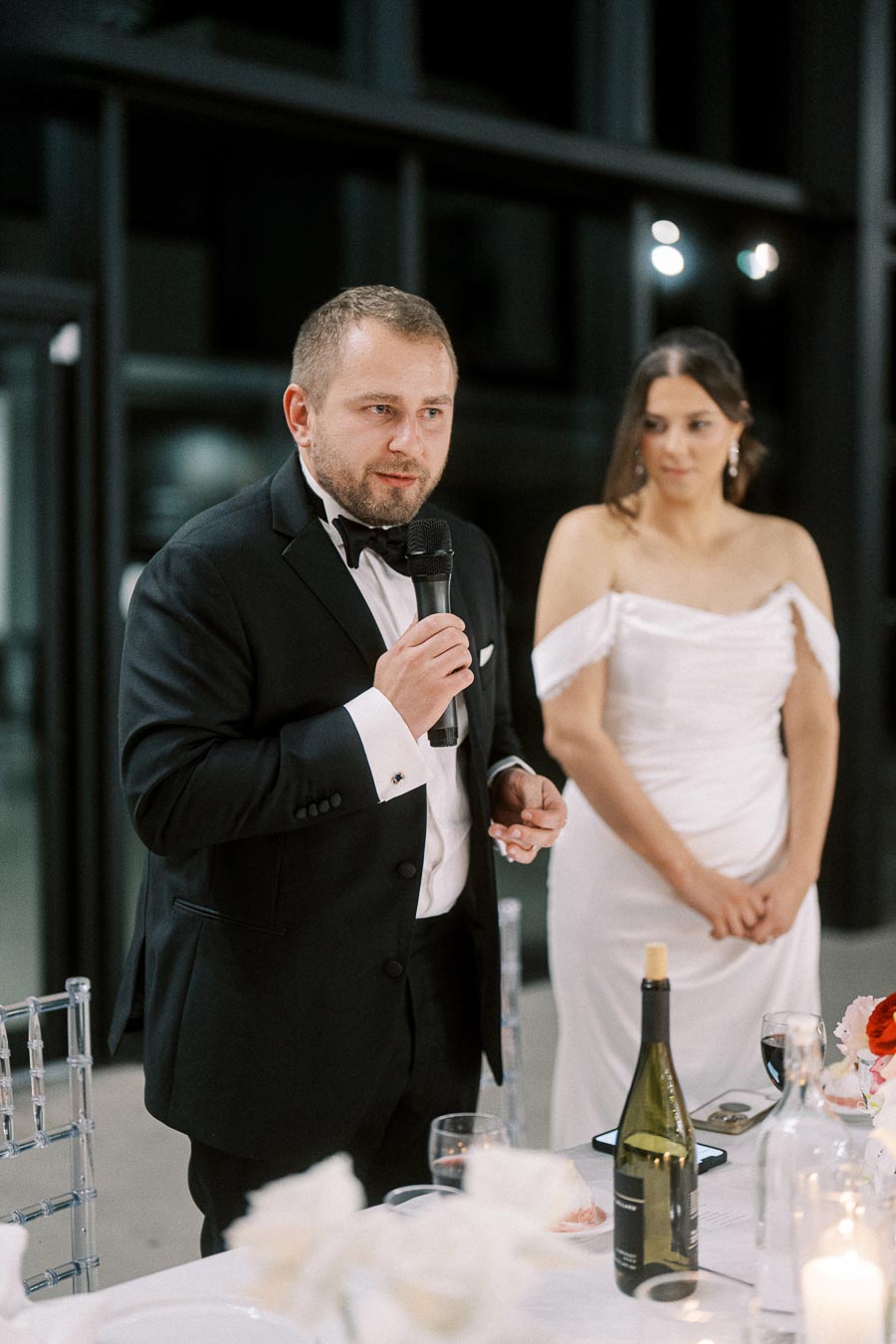 Man giving a speech at a wedding reception, wearing a formal black tuxedo, while a woman in a white dress stands beside him, set in an elegant venue with a decorated table.