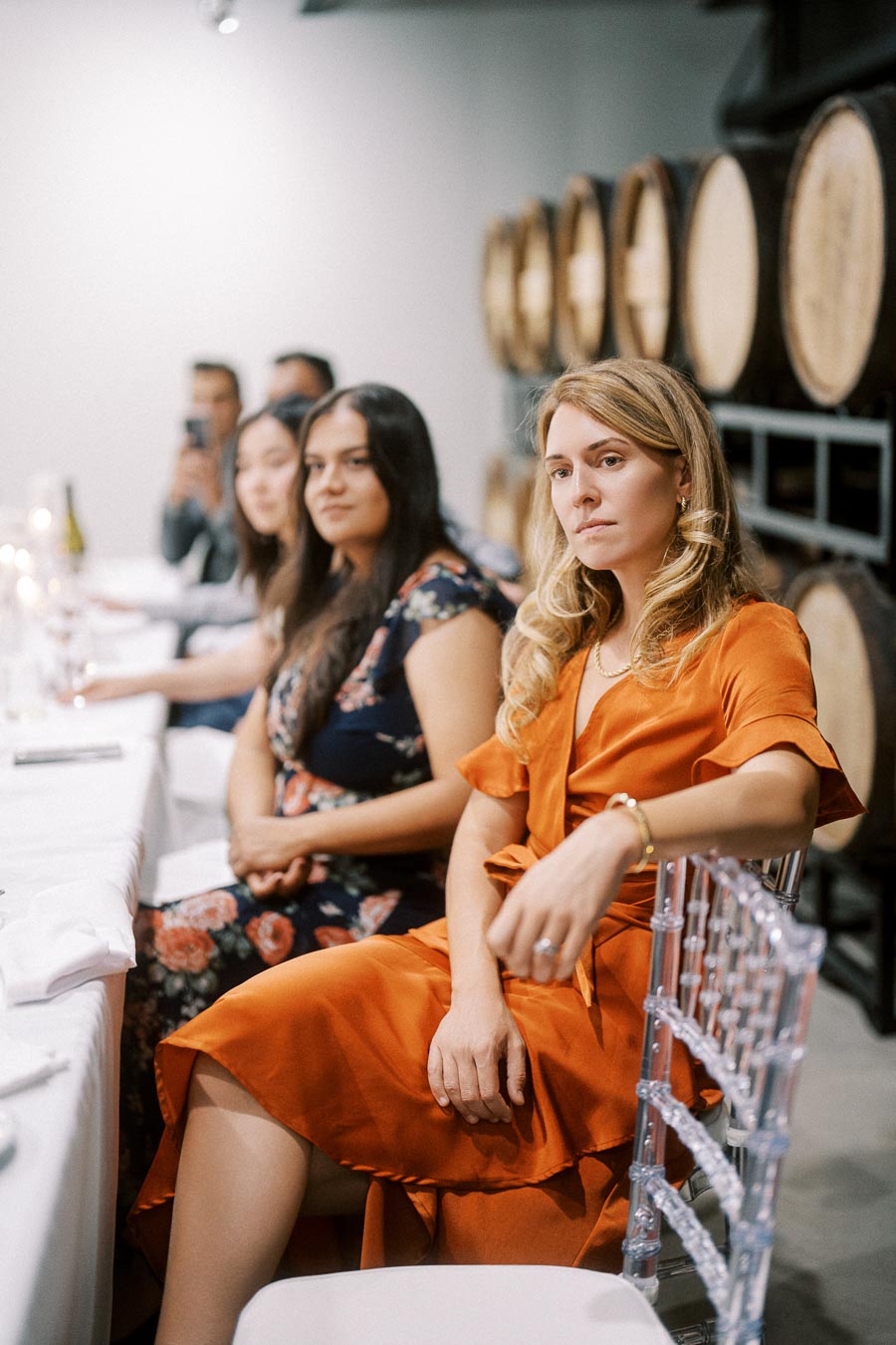 A group of elegantly dressed people seated at a banquet in a winery, with barrels in the background and a woman in an orange dress in focus.