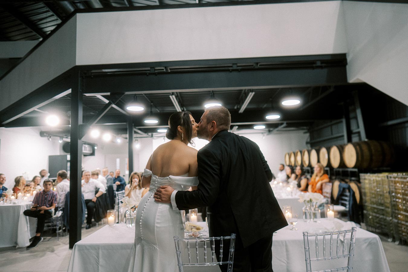 Wedding celebration in modern venue, bride and groom sharing a kiss with guests seated at decorated tables, surrounded by candlelight and barrels in the background.