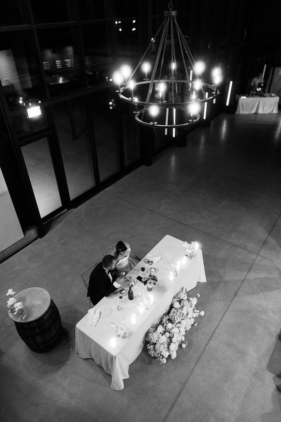 Aerial view of a couple dining at an elegantly decorated wedding reception table, illuminated by a chandelier and surrounded by floral arrangements in a modern event venue.