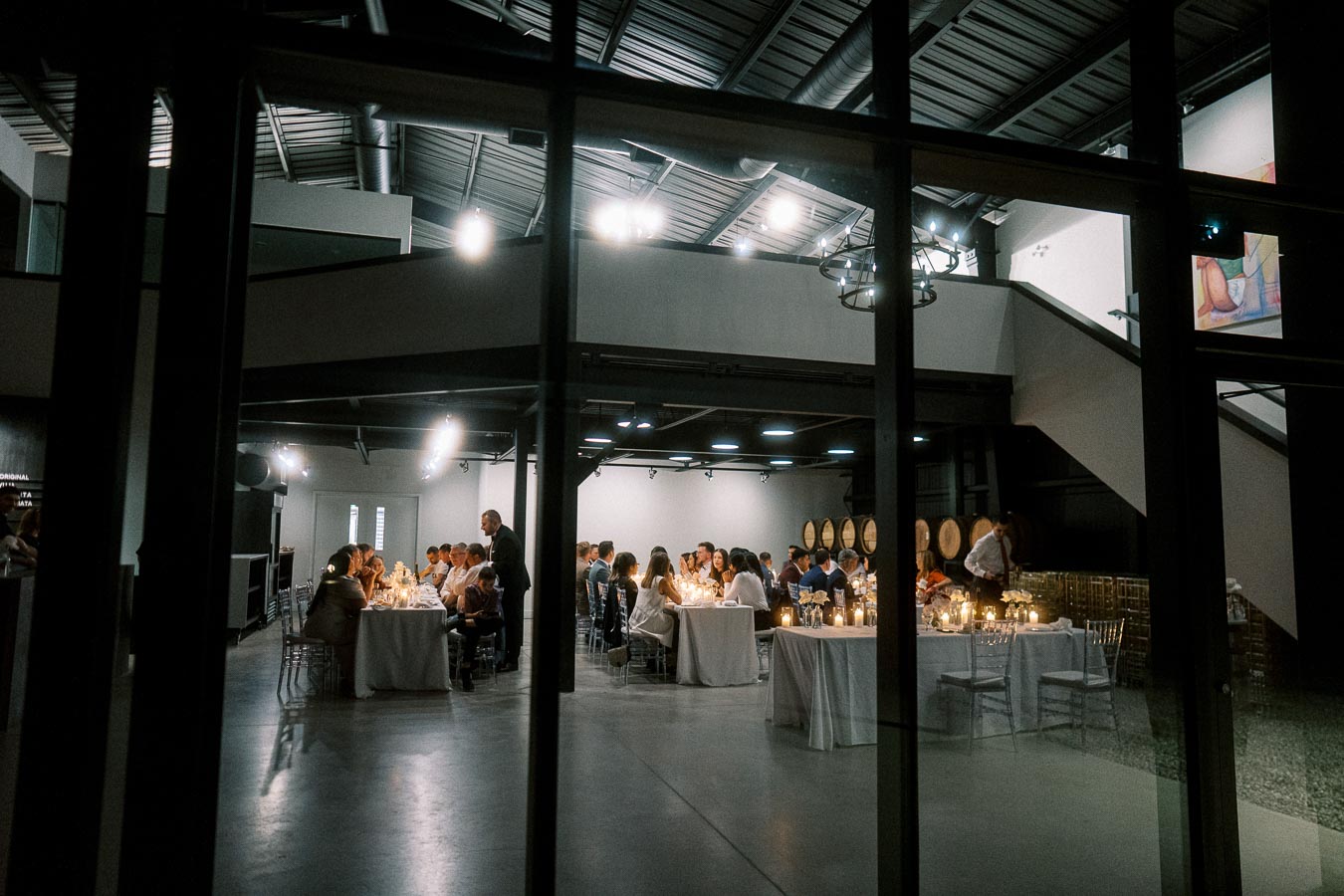Elegant indoor dining event at a modern venue with guests seated at white-clothed tables, softly lit with ambient lighting and surrounded by wine barrels.