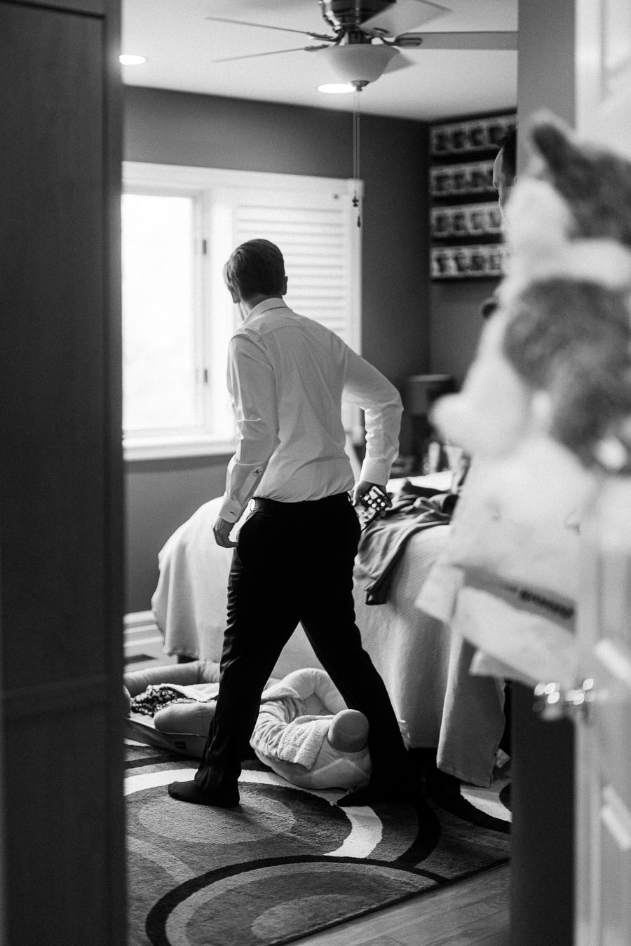 A man in formal attire stands in a bedroom next to a toddler lying on the floor. The room features plush toys, a neatly made