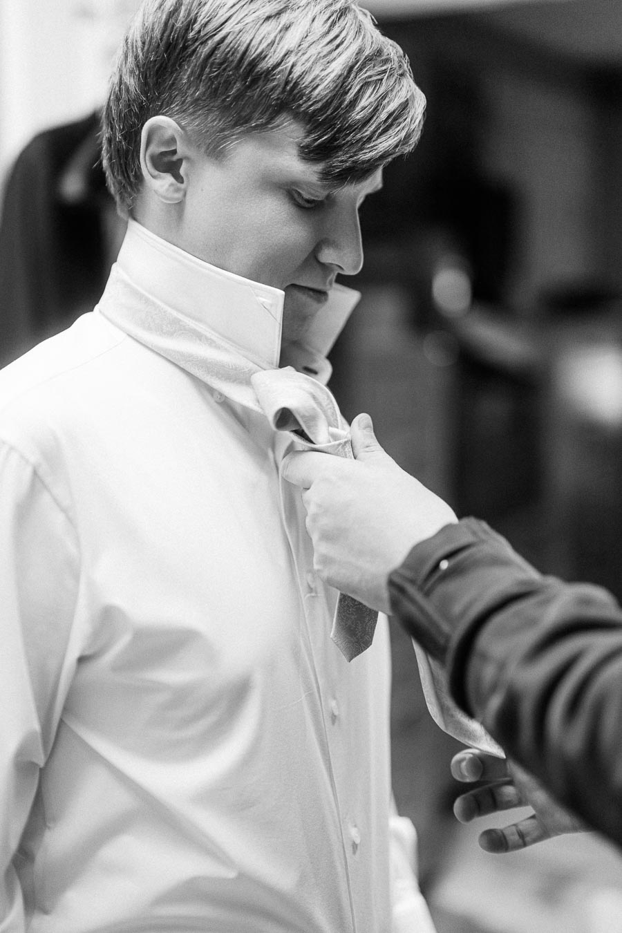 A man is adjusting a tie on a young man in a white dress shirt, preparing him for a formal event in a black and white