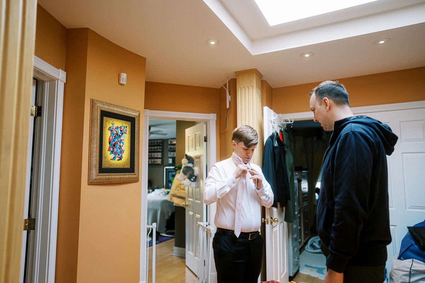 Man in a white shirt adjusting a tie in a warmly lit room, with another person observing, framed artwork on the wall.