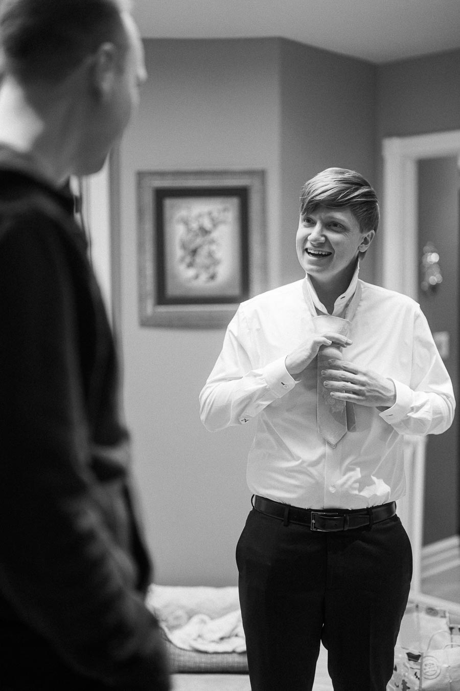 Black and white photo of a young person in a white shirt smiling while adjusting a tie, with another person standing nearby;