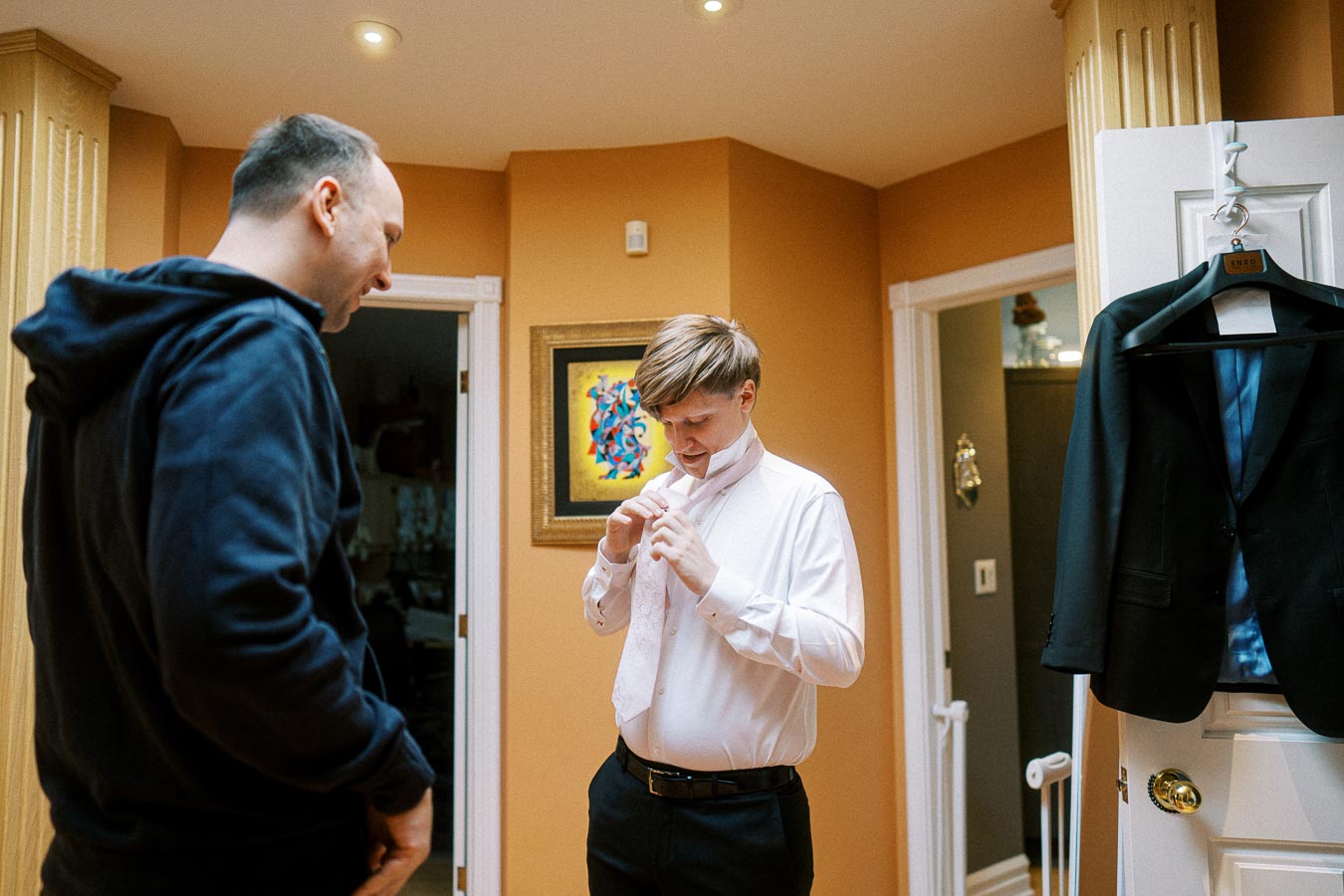 Two men in a warmly-lit room, one adjusting a tie and the other in casual wear. A suit jacket hangs on the door, suggesting