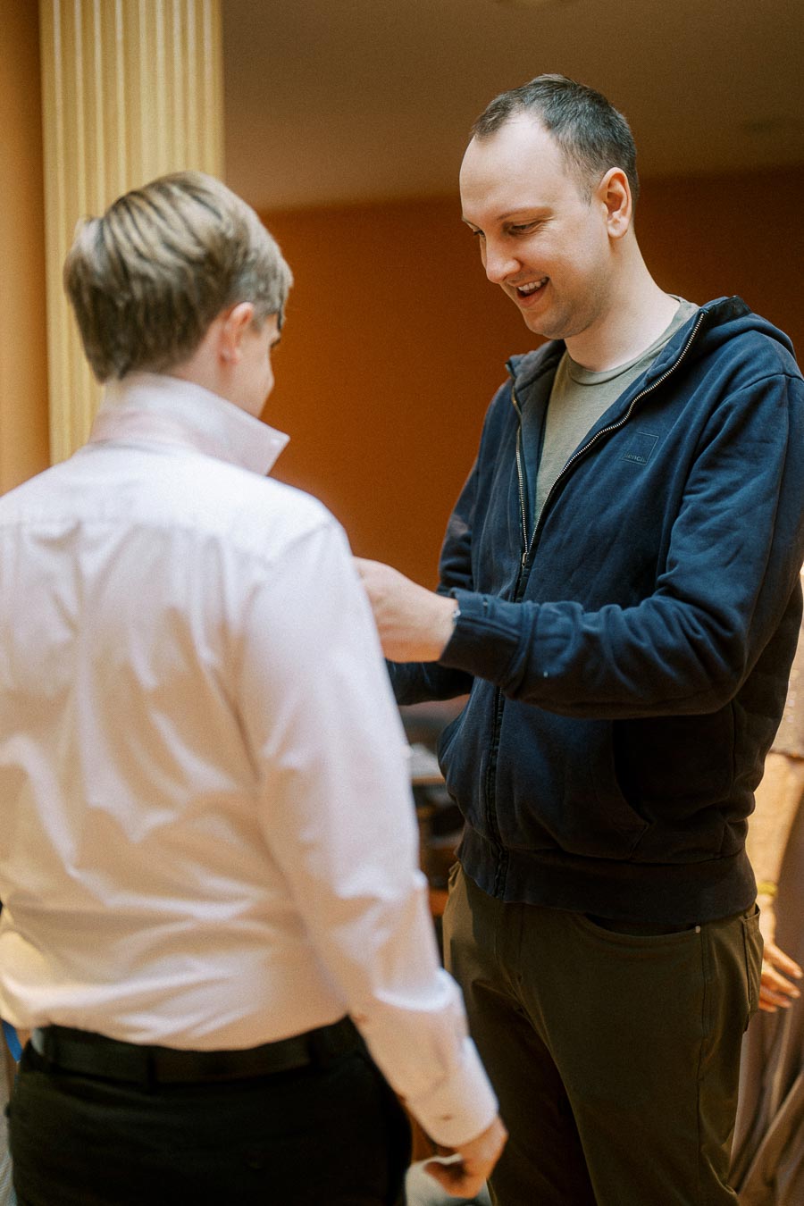 Two men having a conversation indoors, with one wearing a white shirt and the other in a blue hoodie, smiling warmly in a