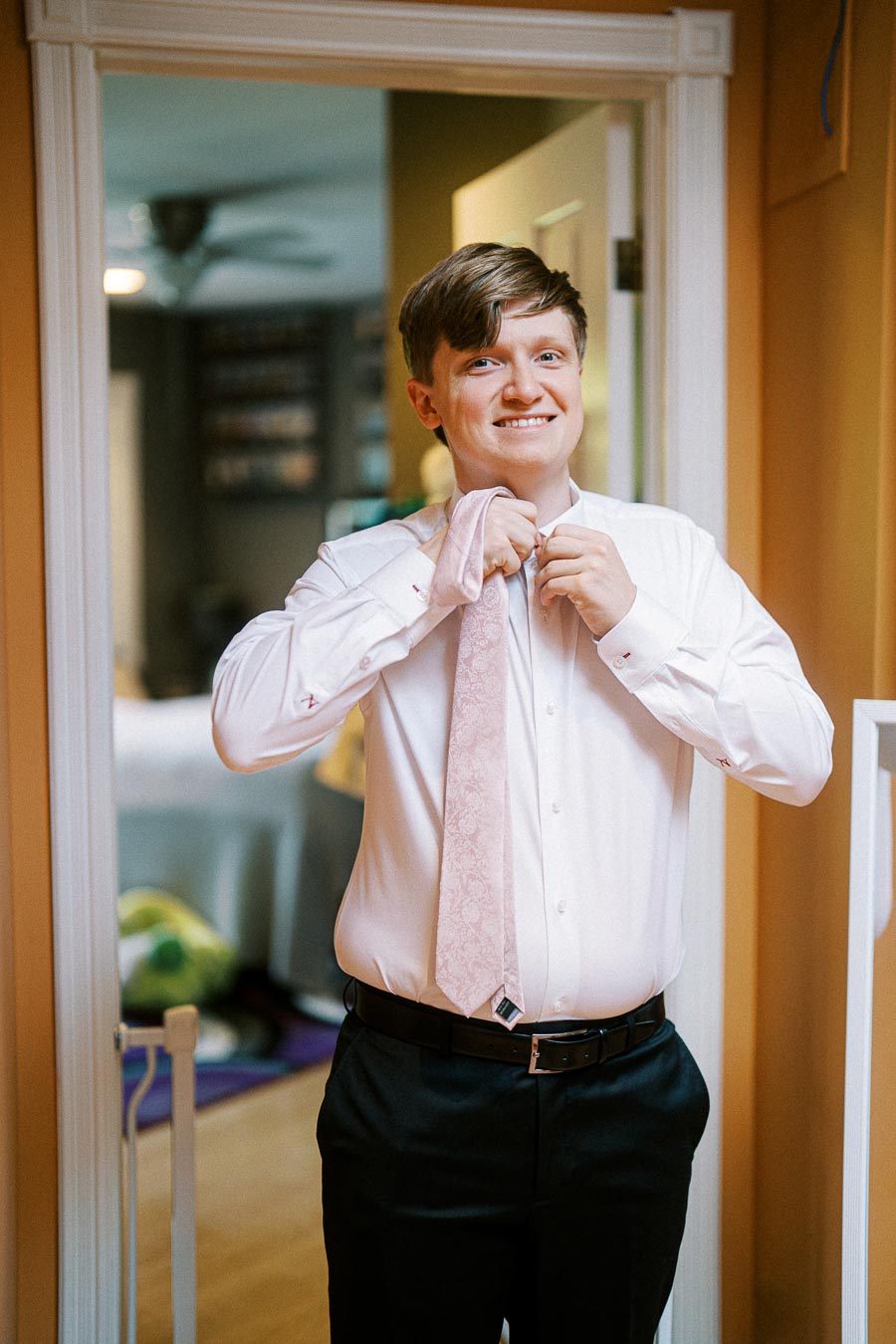 A young man smiling while adjusting his pink tie in a warmly lit room, dressed in a white shirt and black pants.