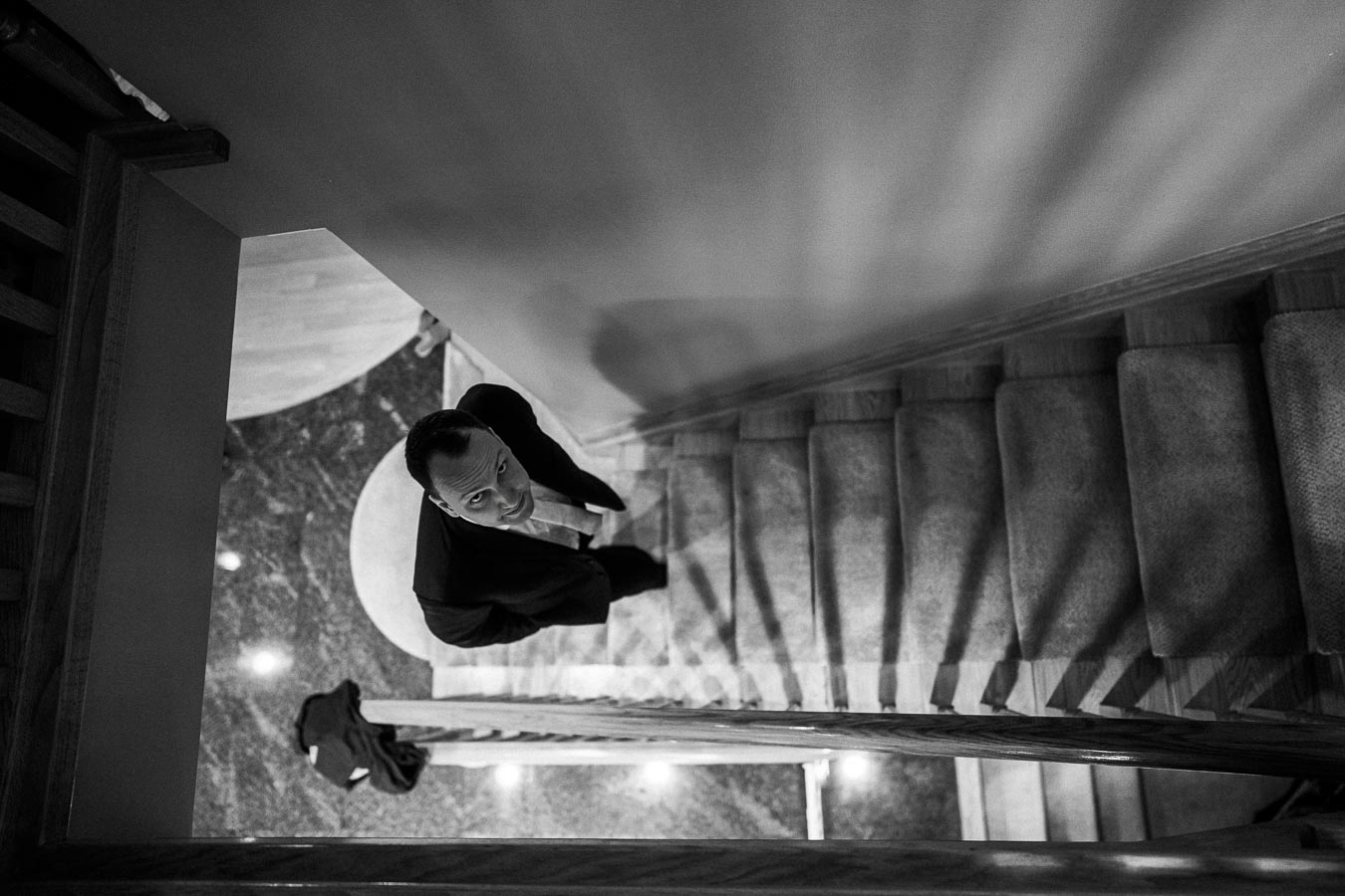 A man in a suit ascending a carpeted staircase, captured from a top-down perspective in black and white, with soft lighting