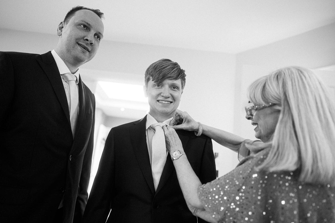 A woman adjusts a young man's tie as he smiles, with another man looking on, all dressed in formal suits, in a well-lit room.