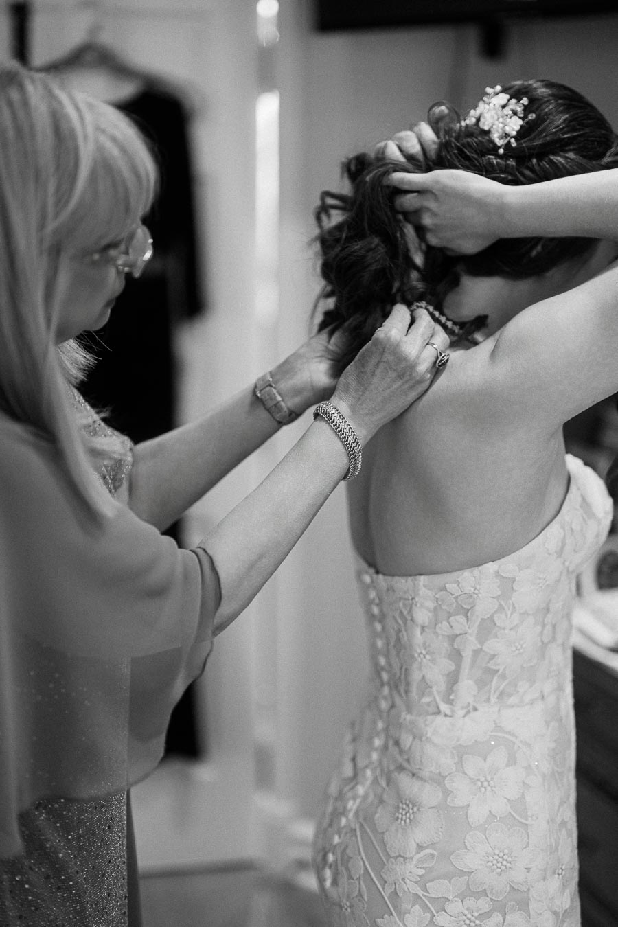 Black and white image of a bride in a floral lace wedding dress being assisted with her necklace by an older woman,