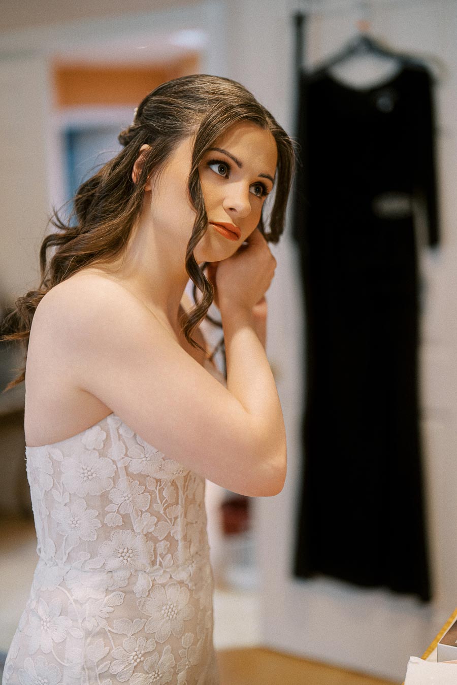 Woman in elegant bridal gown adjusts earring in a softly lit room, with formal black dress hanging in the background.