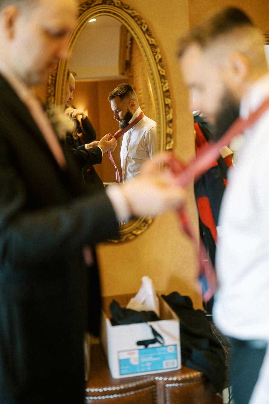 Groom getting ready for wedding, adjusting tie in front of an ornate mirror while wearing formal attire.