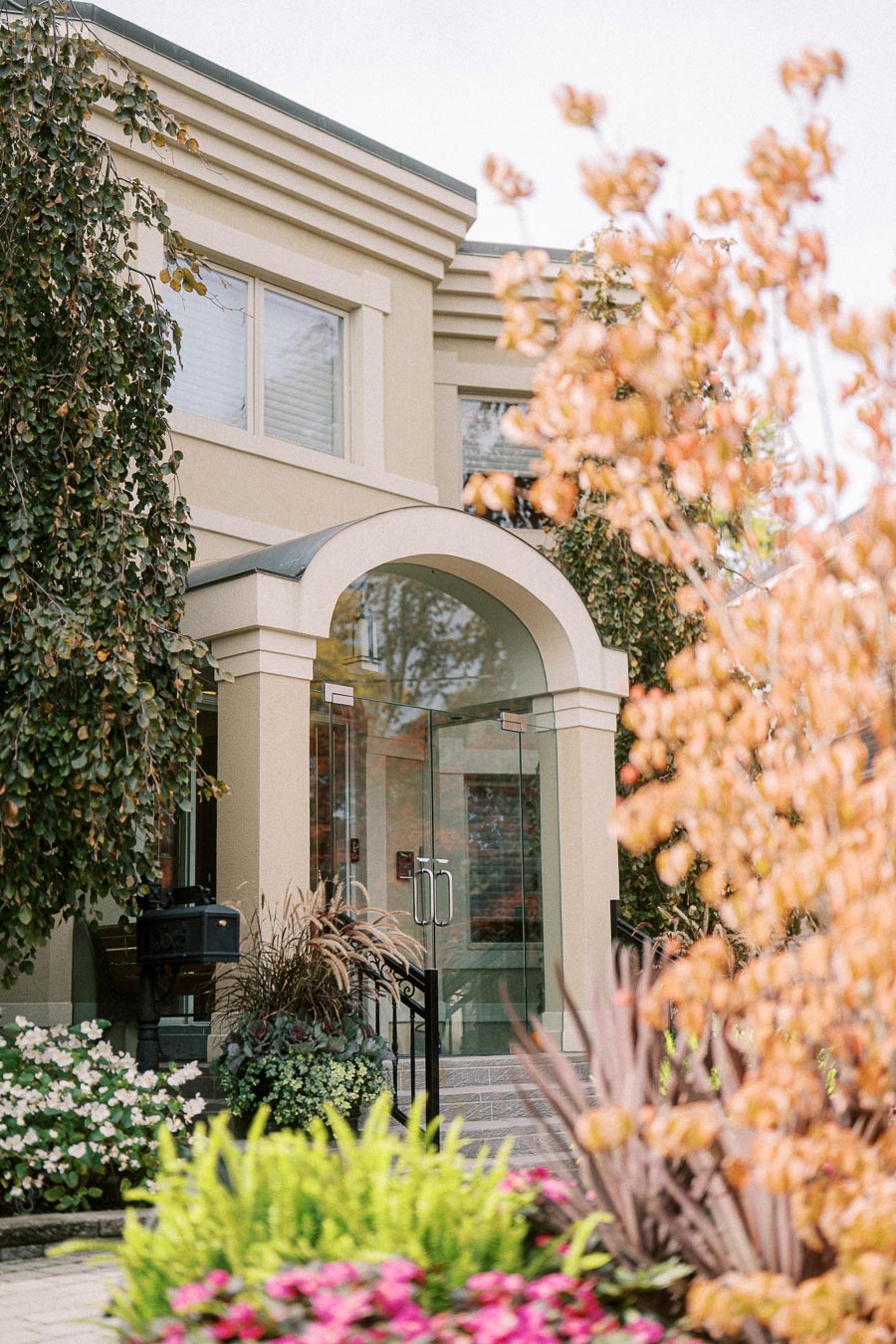 Modern house entrance with glass doors surrounded by lush greenery and colorful flowers in an autumn setting.