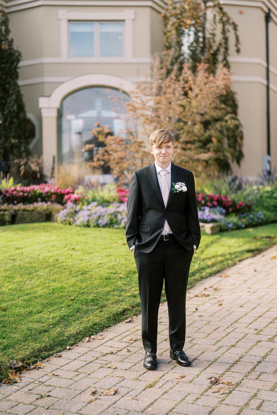 Young man in a suit standing outdoors, posing for a wedding or formal event. The background features a landscaped garden