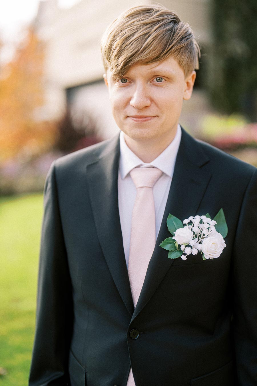 A young man in a black suit with a light pink tie and a white floral boutonnière stands in a garden setting, showcasing a