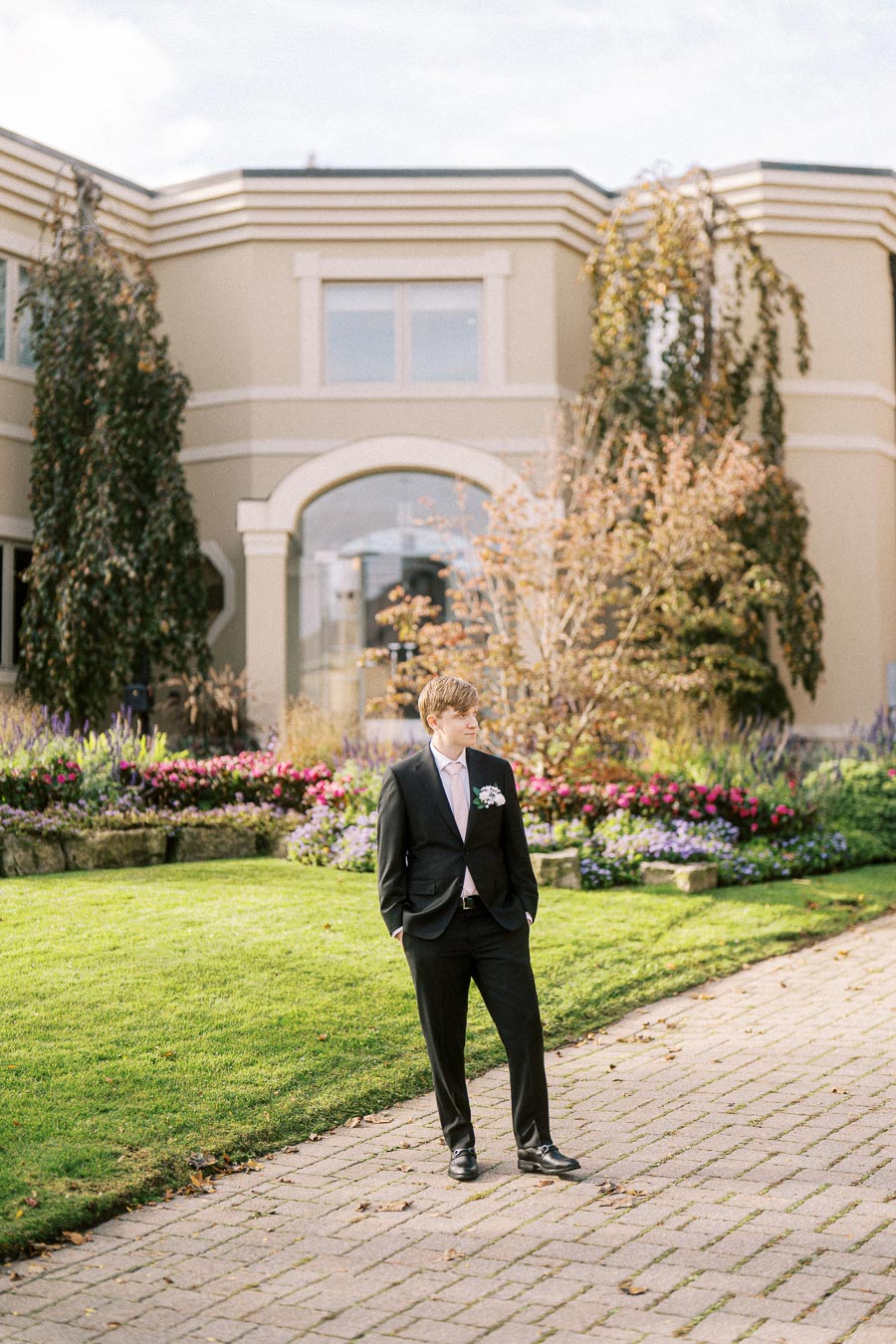 Young man in formal black suit and pink tie standing on a paved path in front of a modern house with lush garden and vibrant