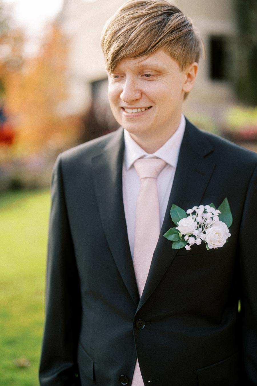 A smiling groom in a black suit with a white rose boutonniere in an outdoor setting.