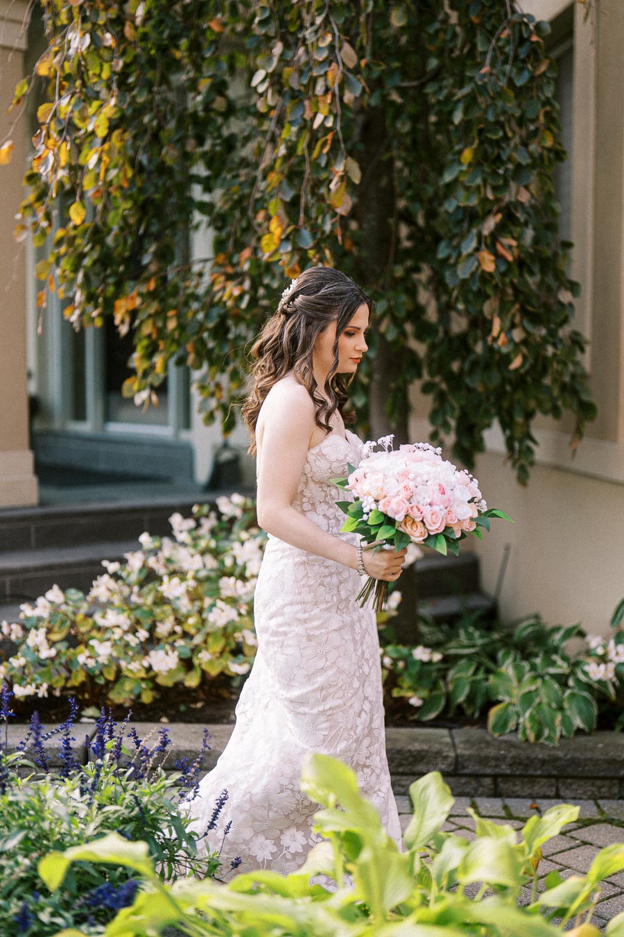 Brunette bride in a lace wedding dress holding a bouquet of pink flowers, walking in a garden setting with lush greenery and