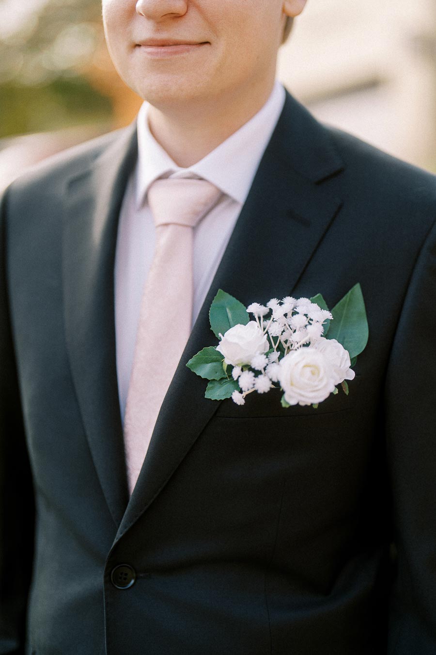 Groom in a black suit with a white boutonniere and pink tie at an outdoor wedding ceremony.