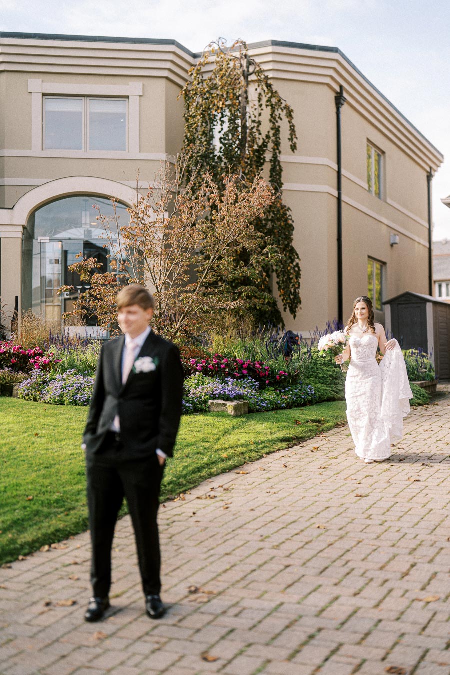 Bride in a white wedding dress walking behind a groom in a black suit during a first look moment in a garden with colorful