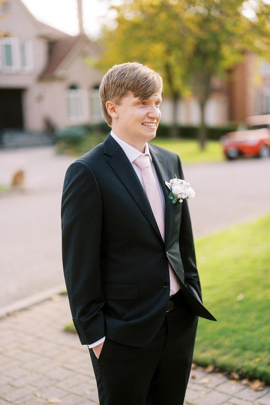 A young man in a suit with a boutonniere smiles outdoors, standing in front of a house and a garden with green grass and