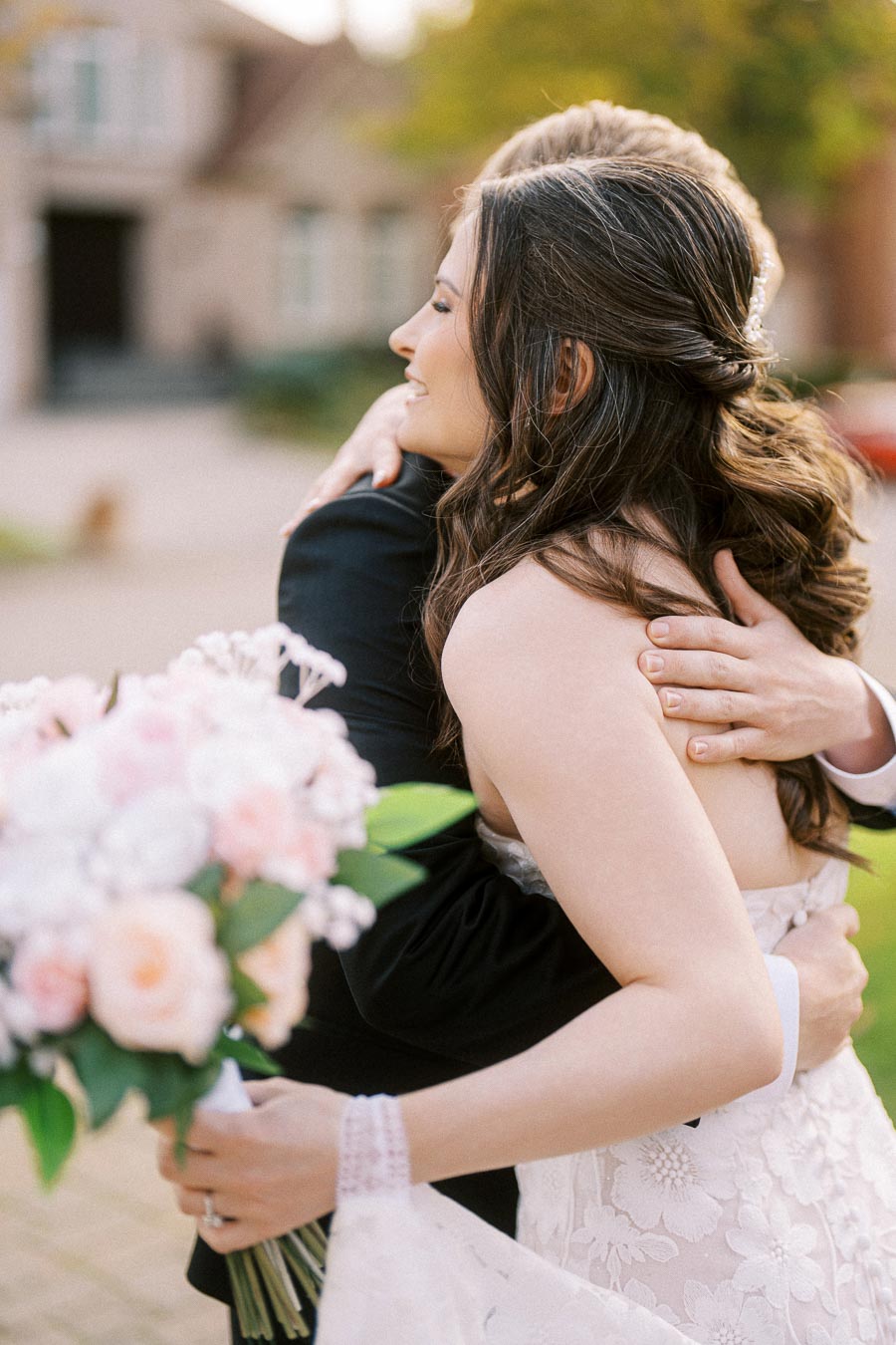 A bride in a white lace dress embraces her partner, holding a bouquet of pink and white flowers, in an outdoor setting with
