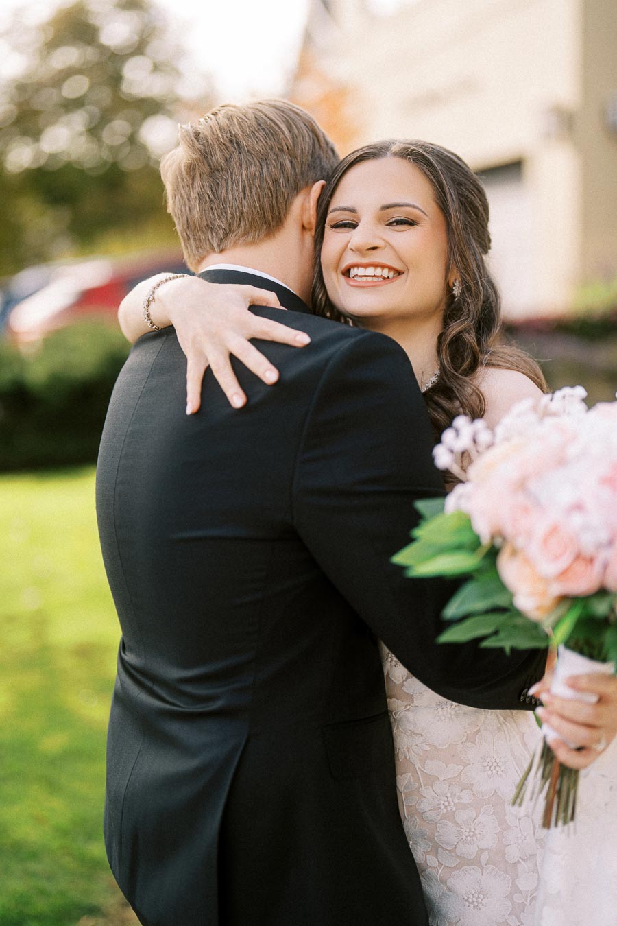 A joyful bride smiling broadly as she embraces the groom, holding a bouquet of pink and white flowers, in a lush green