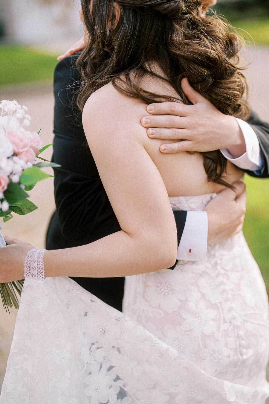 A bride and groom embrace on their wedding day, with the groom in a black suit and the bride wearing a lace wedding dress
