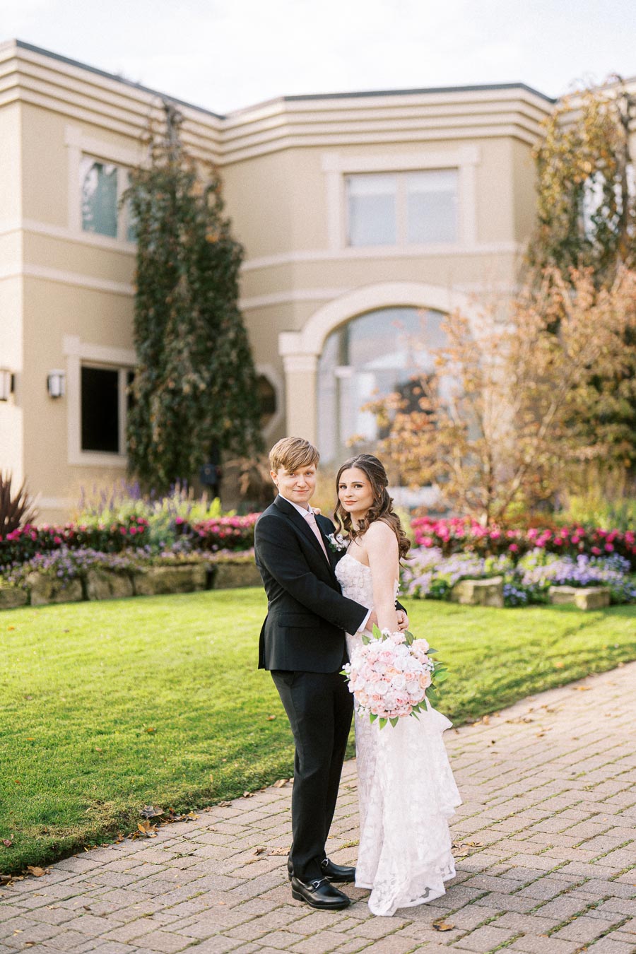 Elegant wedding couple poses in front of a beautifully landscaped garden with blooming flowers and a stylish modern building