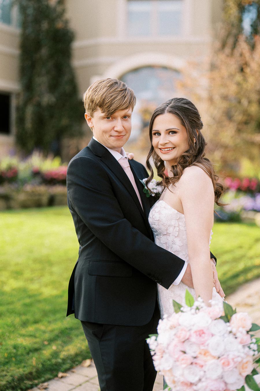 A happy couple embracing on their wedding day in an outdoor garden setting, with the groom in a black suit and pink tie and