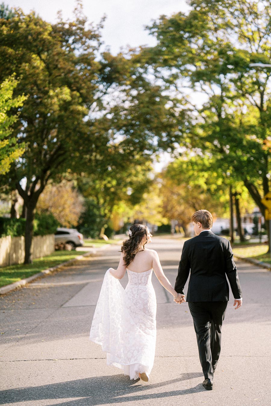 Newlywed couple walking hand in hand on a sunny street, bride in a lace wedding gown and groom in a black suit, with green