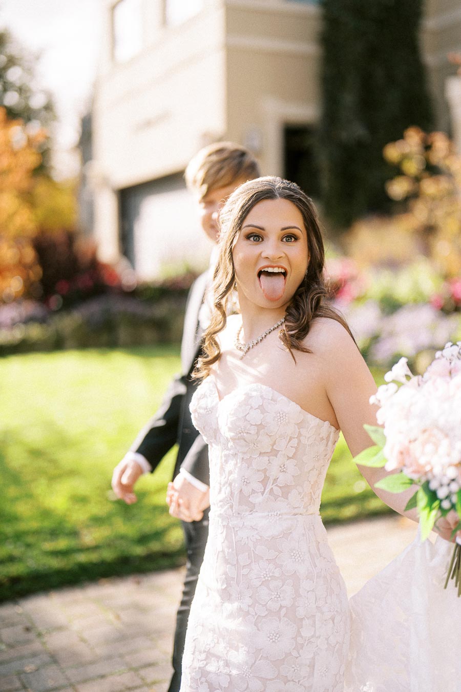 Bride playfully sticking out tongue while walking in garden, wearing a lace wedding dress and holding a bouquet under sunny