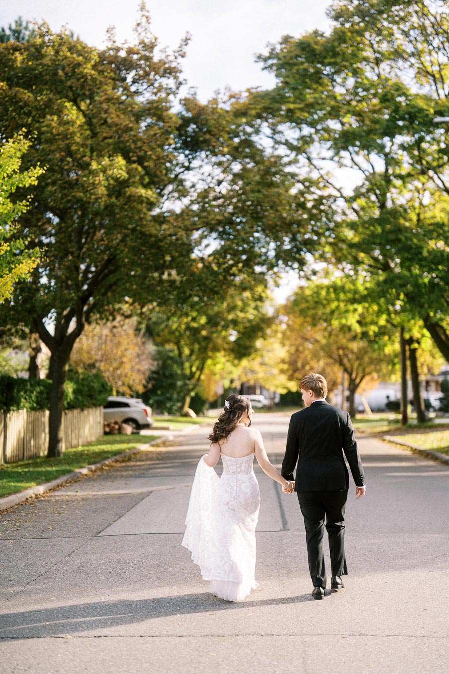 A couple in wedding attire holding hands and walking down a sunlit, tree-lined street in autumn.