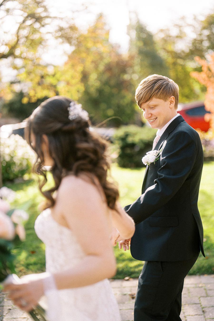 A bride and groom smiling and holding hands, walking outdoors on a sunny day with a vibrant, green garden in the background.