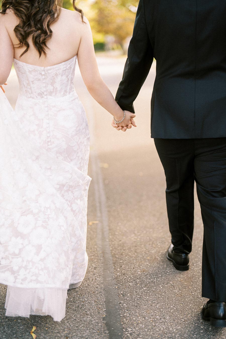 A bride in a flowing white lace wedding dress and a groom in a black suit holding hands as they walk down a sunlit path.