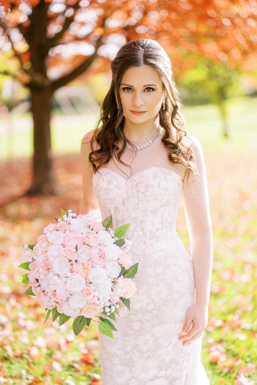 A bride in an elegant white lace wedding dress stands in a sunlit park with autumn foliage, holding a bouquet of pink and