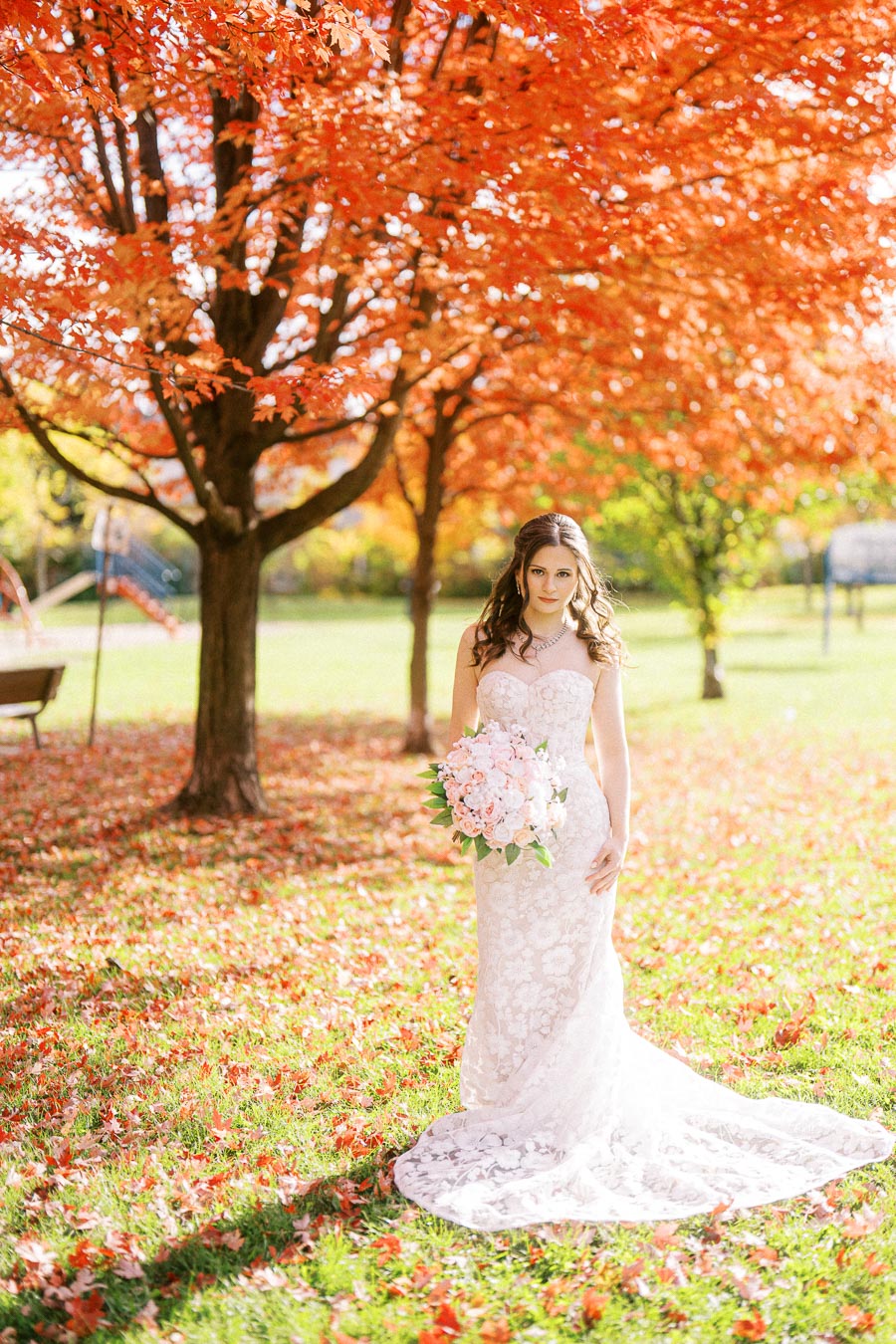 A bride in a lace wedding dress holds a pink bouquet, standing under vibrant red autumn trees on a sunlit day.