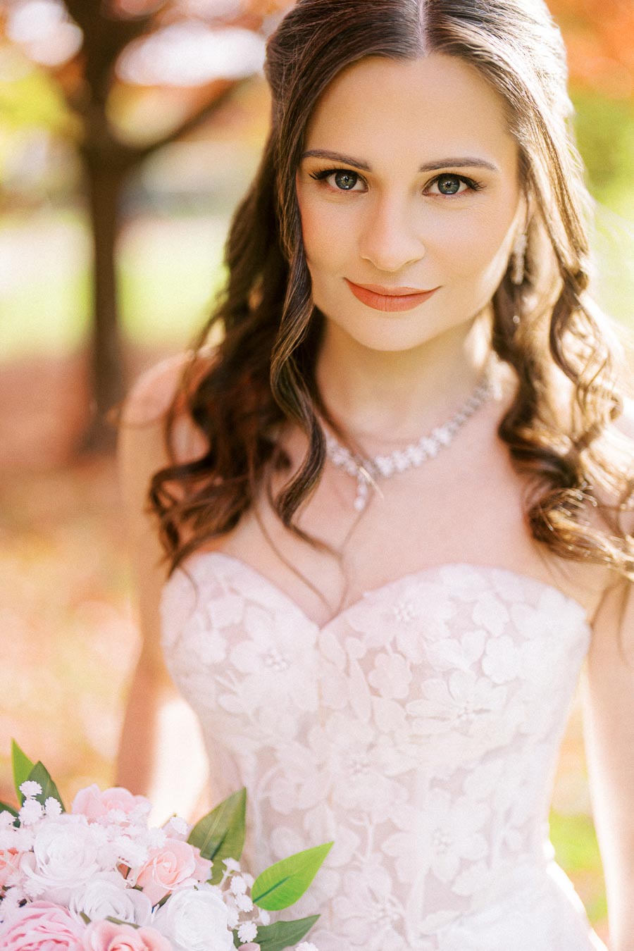 A bride with long brown hair and a strapless lace wedding dress holding a bouquet of pink and white roses, standing outdoors