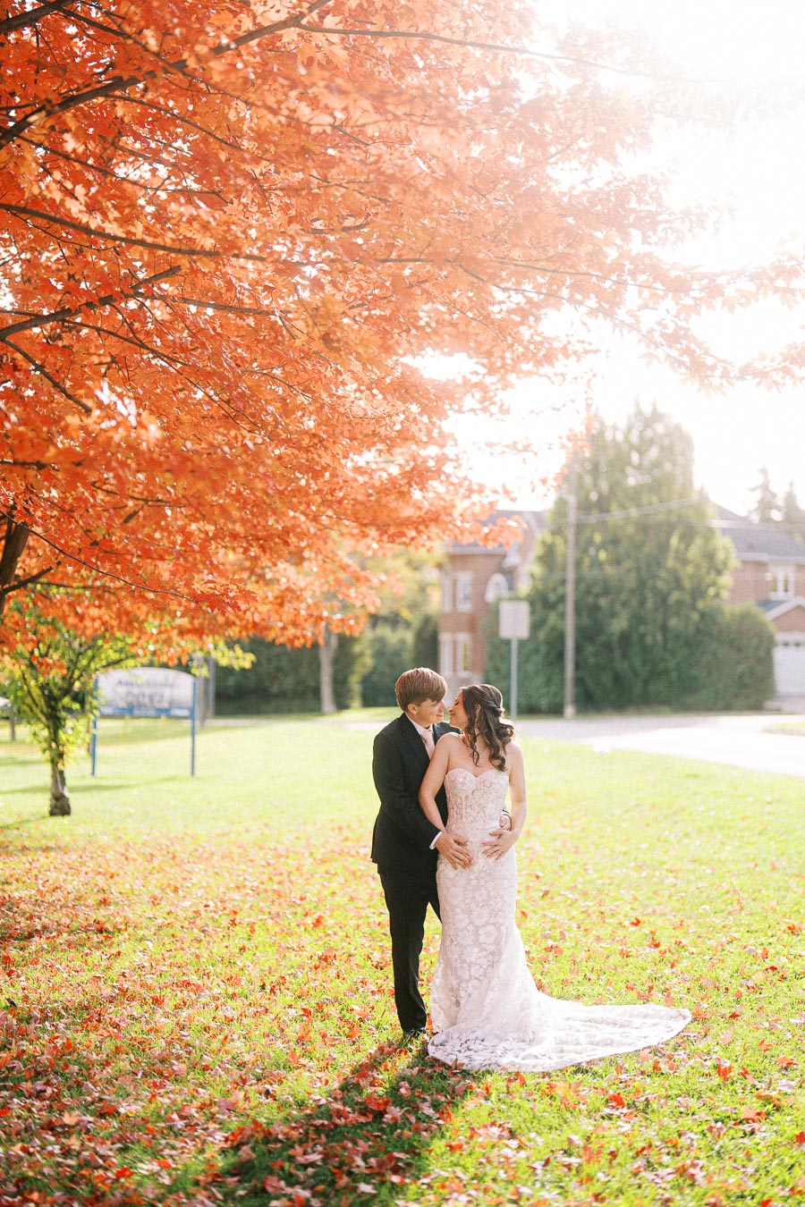 A newlywed couple embraces under a vibrant red autumn tree, surrounded by fallen leaves in a serene park setting, capturing